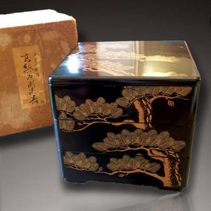 Puede incluir: Una caja lacada japonesa negra de tres niveles con diseños dorados de pinos. La caja tiene forma cuadrada y un acabado brillante. Una caja de cartón con texto japonés es visible en el fondo.