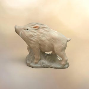 May include: A porcelain figurine of a wild boar standing on a textured base. The boar is colored in soft shades of beige, cream, and light brown, with a smooth, glossy finish. It is depicted in a side profile, showing its snout, ears, and tail.