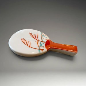 Puede incluir: Un cenicero de cerámica blanca con un mango naranja. El cenicero tiene forma redondeada con un diseño decorativo de líneas naranjas y turquesas. El mango es de color naranja liso.