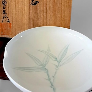 May include: A white ceramic bowl with a delicate bamboo design in shades of green. The bowl has a smooth, glossy finish and a shallow, rounded shape. The bamboo illustration is centered on the bowl's interior.