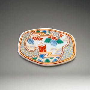 Puede incluir: Un plato decorativo de cerámica de forma ovalada con una base blanca y un diseño colorido e intrincado. El diseño presenta detalles en rojo, azul, verde y dorado, con un motivo central de una estructura roja y follaje estilizado. El plato tiene un borde dorado.