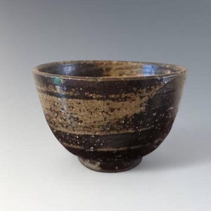 Puede incluir: Cuenco de cerámica artesanal con un diseño a rayas marrón oscuro y beige. El cuenco tiene una forma ligeramente irregular y una base pequeña y redondeada. El interior es oscuro y el exterior presenta un aspecto texturizado y terroso, lo que sugiere una estética rústica.