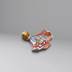 Puede incluir: Un reposapalillos de cerámica con forma de kimono estilizado, que presenta una cabeza de tono dorado brillante y un cuerpo decorado con intrincados patrones geométricos en rojo, azul y verde, motivos florales y finos detalles en dorado.