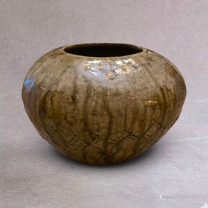 Peut inclure: Un vase en céramique rond et trapu avec une large ouverture et une base effilée. La surface présente un vernis marron et beige tacheté avec des motifs de coulures verticales et une texture gaufrée en losanges sur le bas du corps.