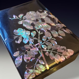 May include: A black rectangular box with a glossy finish, adorned with an iridescent floral design. The design features flowers and leaves inlaid with shimmering, multi-colored mother-of-pearl, creating a striking contrast against the dark background.