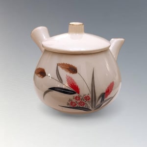 Könnte beinhalten: Cremefarbene Keramik-Teekanne mit Deckel und Ausguss. Die Teekanne ist mit einem Blumendesign mit braunen, roten und schwarzen Akzenten verziert. Die Teekanne hat eine runde Form und einen kleinen Griff auf dem Deckel.