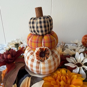 May include: A stack of three fabric pumpkins featuring different patterns. The top pumpkin has a black and beige gingham check, the middle is orange and yellow plaid, and the bottom is orange and white gingham with a central fabric flower and button.