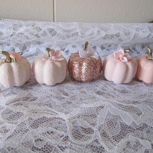 Pode incluir: Cinco ab&oacute;boras decorativas em tons de rosa e ouro rosa. Cada ab&oacute;bora tem um caule dourado e &eacute; adornada com uma flor ou la&ccedil;o. As ab&oacute;boras est&atilde;o dispostas sobre uma superf&iacute;cie de renda branca.