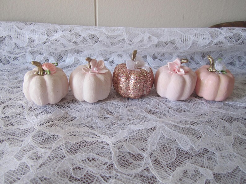 Pode incluir: Cinco ab&oacute;boras decorativas em tons de rosa e ouro rosa. Cada ab&oacute;bora tem um caule dourado e &eacute; adornada com uma flor ou la&ccedil;o. As ab&oacute;boras est&atilde;o dispostas sobre uma superf&iacute;cie de renda branca.