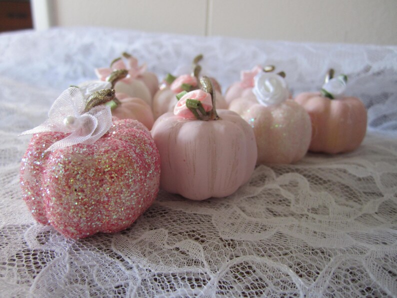 Pode incluir: Uma cole&ccedil;&atilde;o de pequenas ab&oacute;boras decorativas em v&aacute;rios tons de rosa. Algumas s&atilde;o cobertas com glitter, outras t&ecirc;m um acabamento liso e algumas s&atilde;o adornadas com pequenas flores e la&ccedil;os. As ab&oacute;boras est&atilde;o dispostas sobre uma superf&iacute;cie de renda branca.