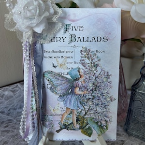 May include: A decorative card titled "Five Fairy Ballads" featuring a fairy illustration with a lilac bush. The card is propped on a small easel and adorned with white lace, pearls, and ribbons. A white rose and other flowers are in the background.