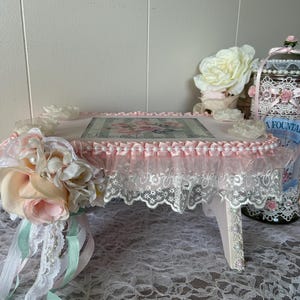 May include: A decorative pink and white serving tray with a floral design on top. The tray is trimmed with lace and pearls, and adorned with fabric flowers and ribbons. The tray has small legs.