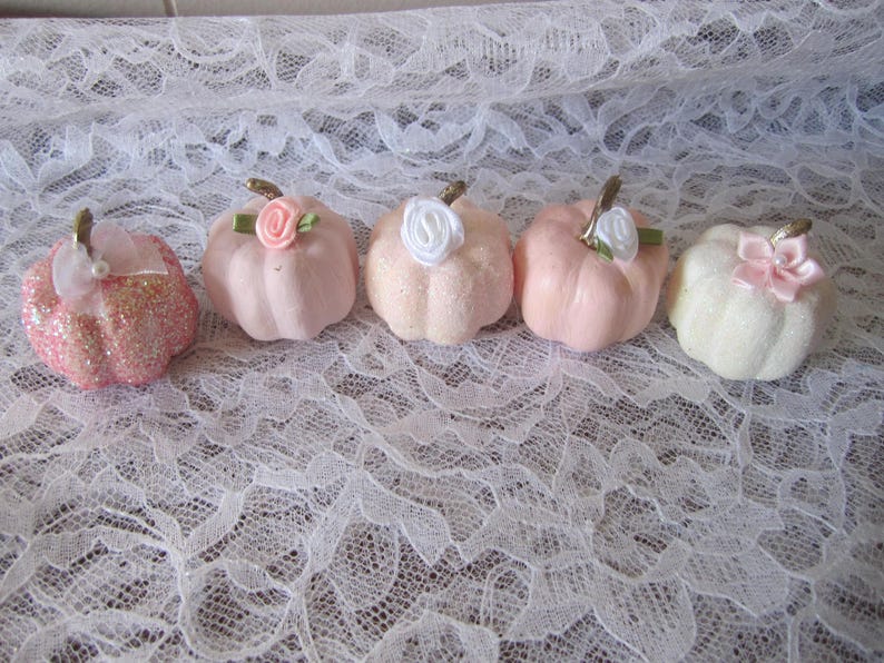 Pode incluir: Cinco ab&oacute;boras decorativas em tons de rosa e branco, cada uma com um caule dourado e uma flor ou la&ccedil;o de tecido. Uma ab&oacute;bora &eacute; coberta com glitter rosa. As ab&oacute;boras est&atilde;o dispostas sobre uma superf&iacute;cie de renda branca.