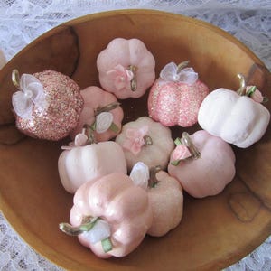 Pode incluir: Uma tigela de madeira cheia de ab&oacute;boras decorativas em v&aacute;rios tons de rosa. Algumas ab&oacute;boras s&atilde;o cobertas com glitter, outras t&ecirc;m la&ccedil;os de tecido e detalhes florais. As ab&oacute;boras t&ecirc;m caules dourados e est&atilde;o dispostas sobre uma superf&iacute;cie de renda branca.