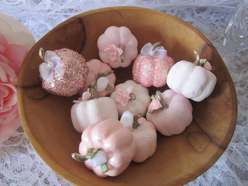 Pode incluir: Uma tigela de madeira cheia de ab&oacute;boras decorativas em v&aacute;rios tons de rosa. Algumas ab&oacute;boras s&atilde;o cobertas com glitter, outras t&ecirc;m la&ccedil;os de tecido e detalhes florais. As ab&oacute;boras t&ecirc;m caules dourados e est&atilde;o dispostas sobre uma superf&iacute;cie de renda branca.