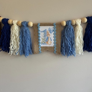 May include: A banner with blue, white, and navy tassels and wooden beads. The center features a burlap pennant with a blue scalloped border and a picture of a bunny holding a balloon with the number 1.