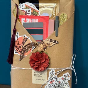 May include: A brown paper-wrapped gift tied with white twine, adorned with stickers, a red flower, and a tassel. The gift contains various items, including a pen, stickers, and a packet of Equal. The quote "Perseverance, secret of all triumphs" is visible.