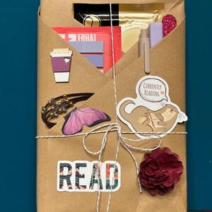 Könnte beinhalten: Ein Geschenk verpackt in braunem Papier, mit weißer Schnur gebunden. Verziert mit Aufklebern mit der Aufschrift "READ" und "Currently Reading", einer lila Kaffeetasse, einem Schmetterling und einer roten Blume. Enthält einen Stift, ein Lesezeichen und andere kleine Gegenstände.