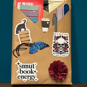 Puede incluir: Un paquete envuelto en papel marrón decorado con pegatinas y pequeños objetos. Las pegatinas incluyen un gato negro sobre libros, una mariposa azul, una calavera con rosas y el texto "smut book energy" y "If it isn't smut I won't read it".