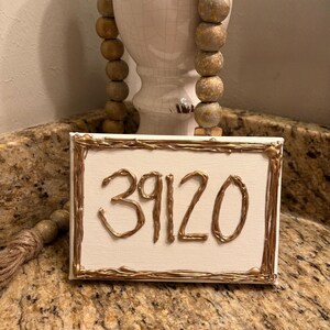 Op de afbeelding: Een rechthoekig wit canvasbord met het getal 39120 geschreven in reliëf metallic goudkleurige verf. Het canvas is omrand door een bijpassende metallic goudkleurige geschilderde lijst, wat zorgt voor een textuurrijk, driedimensionaal uiterlijk.