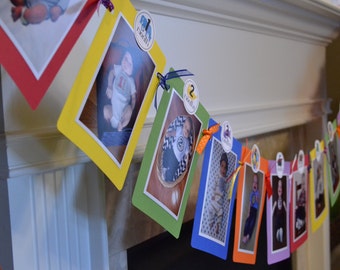 May include: A colorful banner with photos of a baby at different ages. Each photo is framed in a different color and has a number and animal sticker on it. The banner is hanging on a white fireplace mantel.