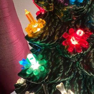 May include: A ceramic Christmas tree with multi-colored lights. The lights are shaped like stars and are lit with clear bulbs. The tree is decorated with a variety of colors, including red, green, blue, yellow, and purple.