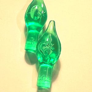 May include: Two green Christmas light bulbs with a starburst design. The bulbs have a teardrop shape and are made of transparent green glass. The bulbs are lit and casting a shadow on a light yellow background.