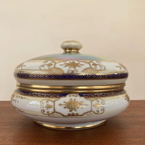 May include: A round porcelain lidded dish featuring a white base with gold floral patterns and a vibrant cobalt blue band. The lid has a central knob and an iridescent finish, while the edges are trimmed with gold accents throughout the piece.