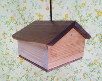 Può includere: Una casetta per uccelli in legno con tetto marrone scuro e corpo marrone chiaro. La casetta è appesa a una corda nera. Il design è una semplice forma di casa, con un corpo rettangolare e un tetto a capanna.