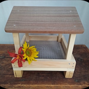 May include: A wooden bird feeder with a rectangular design. The top is a darker wood, while the frame and base are a lighter wood. A mesh screen forms the base of the feeding area. A yellow sunflower and red flower are placed on the front.