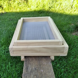 May include: A wooden sifter with a fine mesh screen, resting on a weathered wooden surface. The sifter is made of light-colored wood and is rectangular in shape. The background features green grass and sunlight.