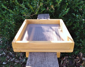 May include: A wooden sifting tray with a fine mesh screen. The tray is made of natural wood and has a rectangular shape. It is sitting on a wooden surface.
