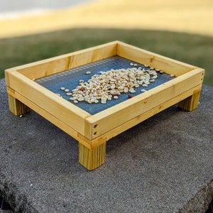 May include: A wooden bird feeder with a wire mesh bottom, filled with peanuts. The feeder is raised on four wooden legs.