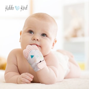 May include: A baby wearing a pink and white teething mitten with a geometric pattern. The mitten has textured silicone bumps for soothing sore gums. The baby is lying on a white blanket.