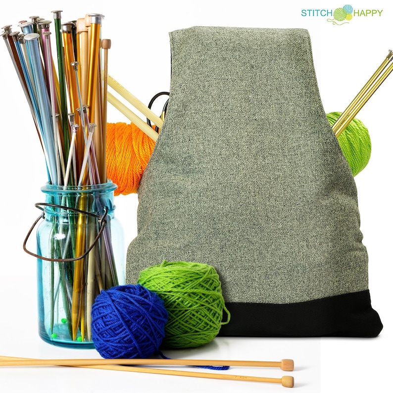 May include: A gray knitting bag with a black bottom, filled with yarn and knitting needles. The bag is sitting on a white surface with two wooden knitting needles in front of it. There are three balls of yarn: one orange, one green, and one blue.