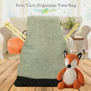 May include: A gray tote bag with a black bottom, labeled "Arm Yarn Organizer Tote Bag" and "Stitch Happy". The bag is filled with yarn, knitting needles, and a crocheted fox toy.