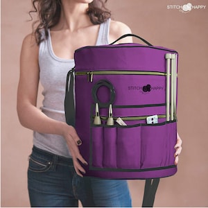 May include: Purple knitting bag with multiple pockets and a zipper closure. The bag has a black strap and the text "Stitch Happy" is embroidered on the front.