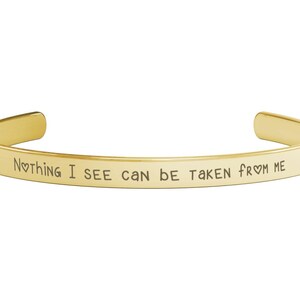 May include: A gold-colored cuff bracelet with the inscription "Nothing I see can be taken from me." The bracelet has a simple, minimalist design and is made of a shiny, polished metal. The text is in a simple, easy-to-read font.