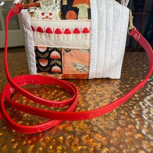 May include: A patchwork crossbody bag with a red strap. The bag features various fabric patterns, including mushrooms, flowers, and geometric designs. The strap is a vibrant red, and the bag has a zipper closure and a small handle.