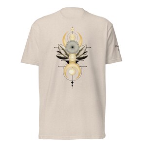May include: A beige short-sleeve t-shirt featuring a central graphic design of a black and gold geometric pattern with leaves, crescent moons, and circular elements. The design is vertically aligned and includes thin black lines and dots.