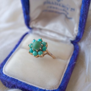 Pode incluir: Um anel de turquesa e ouro com um design de flor. O anel está em uma caixa de anel de veludo azul.