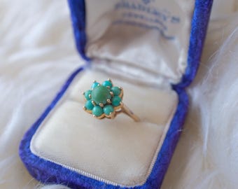 Puede incluir: Un anillo de turquesa y oro con un diseño de flor. El anillo está en una caja de anillo de terciopelo azul.