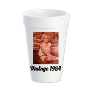 May include: White foam cup with a photo of a young girl in a yellow and white dress sitting on a wooden bench. The text "Vintage 1984" is printed below the photo.
