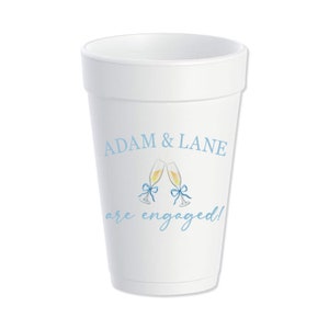 May include: White foam cup with the text "Adam & Lane are engaged!" in blue lettering. Two champagne flutes with blue bows are pictured above the text.