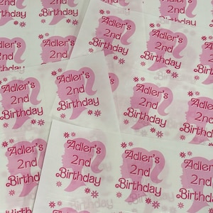 May include: Pink and white favor tags with a silhouette of a woman's head and the text "Adler's 2nd Birthday" repeated on each tag.