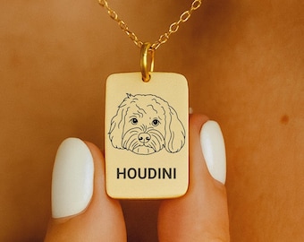 May include: A gold-colored rectangular pendant necklace with a small dog illustration and the name "HOUDINI" engraved below the dog's image. The pendant is held by a person with white nail polish.