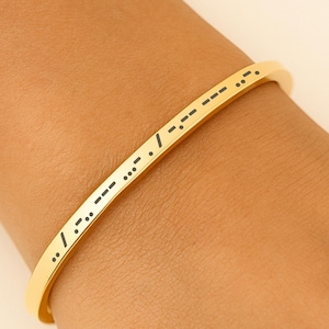 May include: A gold-colored bangle bracelet with black Morse code symbols engraved on the surface. The bracelet is worn on a wrist with white nail polish.