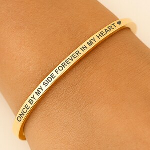 May include: A gold-colored bangle bracelet with the inscription "ONCE BY MY SIDE FOREVER IN MY HEART" in a delicate font. The bracelet is worn on a wrist, showcasing its elegant design and message. The bracelet has a heart symbol at the end of the text.