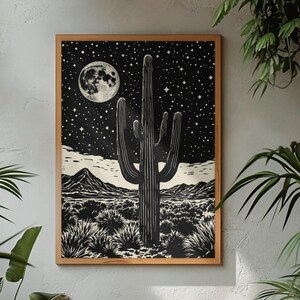 May include: Black and white framed art print depicting a desert scene with a cactus under a starry night sky and a full moon. The artwork is in a wooden frame and is displayed on a grey wall, surrounded by plants.