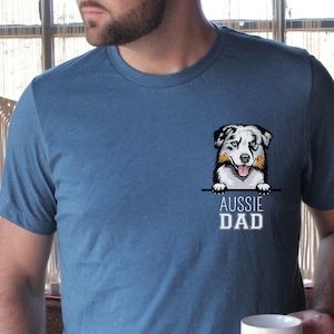 May include: A man wearing a blue t-shirt with a cartoon Australian Shepherd peeking over the top of a pocket. The text "Aussie Dad" is printed below the dog.